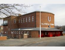 Backsteinbau der U-Bahn-Station "Olympia-Stadion" in Berlin mit geschwungener Fassade, Schriftzug und Uhr über dem Eingang. Im Vordergrund sind rote Türen und ein gepflasterter Vorplatz zu sehen, umgeben von kahlen Bäumen.
