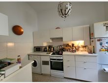 Kleine Küche mit weißen Einbauschränken, Herd, Backofen, Waschmaschine und Kühlschrank. Der Arbeitsbereich ist mit Kochutensilien und Gewürzen gefüllt, eine Lampe leuchtet die Arbeitsfläche aus, und eine moderne Hängelampe befindet sich an der Decke.