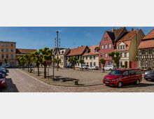 Kleiner Marktplatz mit gepflastertem Boden, umgeben von historischen Häusern mit Fachwerk- und Putzfassaden, sowie kleinen Bäumen in symmetrischen Reihen. Im Vordergrund parken Autos, darunter ein rotes Fahrzeug, unter einem klaren, blauen Himmel.