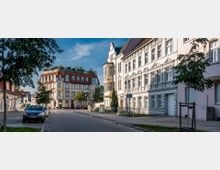 Straßenszene in einer Stadt mit historischen Gebäuden, darunter ein helles Gebäude mit verzierten Fassaden und rechts daneben ein rotes, mehrstöckiges Haus mit Dachbegrünung. Im Vordergrund sind Bäume, parkende Autos und ein Gehweg zu sehen.