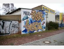 Außenansicht einer Gebäudewand mit auffälligem Graffiti, das bunte Schriftzüge und Autos als Motive zeigt. Daneben eine gemauerte Wand mit weiteren Graffitis; im Hintergrund ist eine Werkstatt mit der Aufschrift "Reifen-Müller" zu sehen, angrenzend an einen gepflasterten Gehweg.