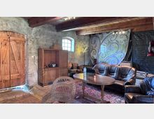 Rustikaler Innenraum mit groben Steinwänden, sichtbaren Holzbalken und einem großen Holztor neben einem kleinen Rundbogenfenster. Im Vordergrund stehen Ledersofas um einen ovalen Holztisch auf einem Teppich; im Hintergrund sind ein Holzschrank, gestapelte Stühle und eine dekorative bemalte Wand zu sehen.