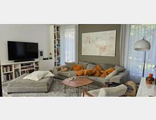 Ein modernes Wohnzimmer mit einer großen grauen Sofalandschaft, dekoriert mit orangefarbenen Kissen, einem Holzcouchtisch und einem hellen Teppich. An der Wand hängt ein abstraktes Kunstwerk, daneben stehen hohe Bücherregale, große Fenster mit weißen Vorhängen lassen viel Tageslicht herein, und ein Wandfernseher ist links angebracht.