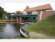Brücke mit grüner Stahlkonstruktion und roten Backsteinpfeilern über einen Fluss, daneben ein Gebäude mit roten Dachziegeln und Fachwerkdesign. Im Vordergrund ein Weg mit Geländer entlang einer begrünten Böschung.