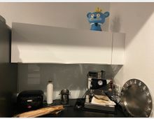 Kleine Küchenzeile mit einer glänzenden Espressomaschine, einer Küchenrolle, einem schwarzen Brotkasten und einem silbernen Tablett auf der Arbeitsfläche. Darüber befindet sich ein weißer Hängeschrank mit einer blauen Figur auf dem Schrank.