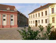 Straßenszene mit historischen Gebäuden, bestehend aus einem hellgelben und einem rosa Gebäude mit Stuckverzierungen und Skulpturen auf dem Dach. Der gepflasterte Platz im Vordergrund ist von etwas Grün und vereinzelten Laternen umgeben.