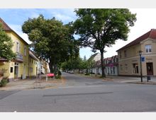 Straßenansicht in einer kleinen Stadt mit niedrigen Häusern, teils gelb oder beige gestrichen, und roten Ziegeldächern. Die Straße ist von Bäumen gesäumt, mit Gehwegen und Geschäftsfronten wie einem Sportgeschäft auf der rechten Seite.