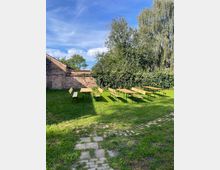 Wiese in einem ummauerten Innenhof mit mehreren Holzbänken und Tischen, links eine alte Backsteinmauer und im Hintergrund dichtes Grün mit hohen Bäumen. Im Vordergrund verläuft ein unregelmäßiger Pflasterweg durch das Gras unter blauem Himmel.