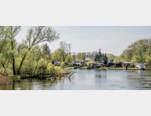 Flusslandschaft mit sanft geschwungenem Ufer, umgeben von Bäumen und Sträuchern, im Hintergrund eine Ansiedlung aus kleinen Häusern und Gärten. Der Fluss spiegelt das Grün der Umgebung wider, und die Szenerie wirkt ruhig und naturbelassen.