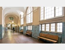 Ein langer, heller Flur mit hohen Fenstern, bogenförmigen Decken und türkisfarbenen Wandfliesen. Entlang der Wände stehen hölzerne Sitzbänke, und Kugel-Deckenleuchten hängen von der Decke.