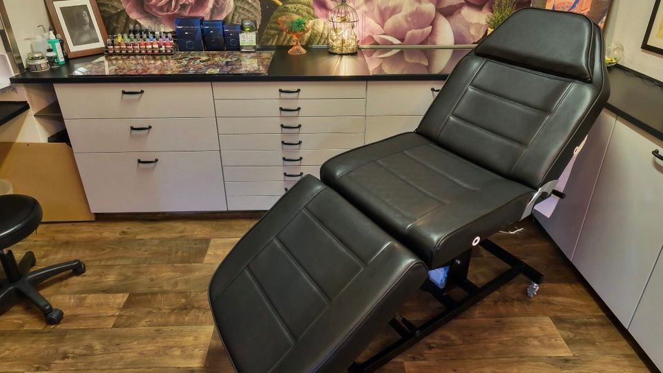 Innenraum eines Tattoo- oder Kosmetikstudios mit einer schwarzen, verstellbaren Behandlungsliege auf Rollen vor einer langen Arbeitsfläche mit vielen Schubladen. An der Rückwand hängt eine großflächige Tapete mit rosa Blumenmotiv; auf der Ablage stehen kleine Fläschchen, Utensilien und gerahmte Bilder.