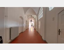 Ein heller, länglicher Flur mit weiß gestrichenen Wänden, roten Böden und einer gewölbten Decke. Auf der rechten Seite sind weiße Türen und Fenster mit Glasbausteinen, während links Heizkörper und Wandnischen zu sehen sind; die Deckenlampen hängen mittig im Gang.