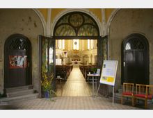 Eingangsbereich einer Kirche mit Buntglasfenstern und zwei offenen Türen, die den Blick auf das Kirchenschiff und einen Altar mit Kreuz freigeben. Links und rechts befinden sich dekorative Elemente, ein Plakatständer und Stühle; der Boden ist mit gemusterten Fliesen bedeckt.