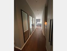 Ein moderner Flur mit dunklem Holzboden sowie drei großen, schmalen Wandspiegeln auf der linken Seite. Am Ende des Flurs ist eine offene Küche mit grauen Schränken und heller Arbeitsplatte zu sehen.