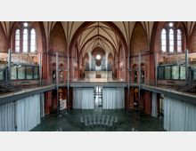 Innenraum einer umgebauten Kirche mit neugotischen Architekturmerkmalen, wie spitzbogigen Gewölben und verzierten Fenstern. Der Raum wurde modernisiert, mit Stahlkonstruktionen, einer Empore, Glasräumen und einer Orgel im oberen Bereich, während im unteren Bereich Sitzstühle in Kreisform sowie große weiße Vorhänge sichtbar sind.