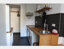 Kleine, schmale Küche mit schwarzen Fliesenwänden, Arbeitsplatten aus Holz und grauem Fliesenboden. Ausstattung umfasst ein Waschbecken, einen Herd mit Backofen, offene Regale und eine Wandhalterung mit Küchenutensilien.