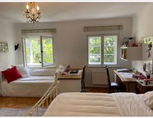 Helles Schlafzimmer mit Holzboden, zwei Fenstern mit Blick auf grüne Bäume, einem Einzelbett mit weißen Decken und rotem Kissen, einem Schreibtisch mit Stuhl und Regalen sowie einem Kronleuchter an der Decke.