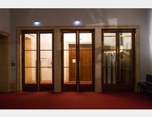 Eingangsbereich eines Gebäudes mit drei großen Türen aus Holz und Glas, eingerahmt von hellem Stein. Der Boden ist mit rotem Teppich ausgelegt, und über den Türen befinden sich beleuchtete Notausgangsschilder.