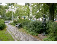 Ein kleiner Park mit einem gepflasterten Weg, einer Holzbank und einer Bronzestatue einer stehenden menschlichen Figur. Im Hintergrund sind Bäume, Büsche, ein kunstvolles Windspiel und Wohnhäuser sichtbar.