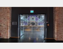 Blick durch eine geöffnete Glastür in eine Veranstaltungshalle mit modernen, violett beleuchteten Dachbalken und einer großen Theke im Vordergrund. Wände aus Ziegelstein, polierter schwarzer Boden und ein Notausgangsschild über der Tür ergänzen die Szene.