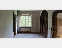 Ein leerer Raum mit einem großen Fenster, das einen Blick auf eine grüne Landschaft bietet. Auf der rechten Seite befindet sich eine doppelflügelige Holztür mit abgerundeten Bögen, und rechts daneben ist ein freiliegendes Mauerwerk sichtbar, das einen Übergang zu einem anderen Raum markiert.