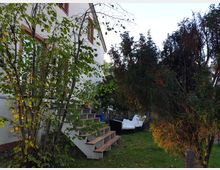 Ein Haus mit weißer Fassade und roten Fensterrahmen, umgeben von Bäumen und Sträuchern. Im Vordergrund befindet sich eine Treppe aus Holzstufen sowie eine Grünfläche mit Gartenmöbeln, darunter ein Sofa in der rechten Bildhälfte.