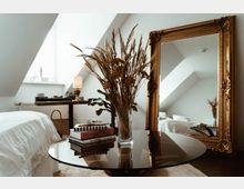 Schlafzimmer mit Dachschrägen, einem runden Glastisch mit Trockenblumen in einer Vase, einem Stapel Bücher und einem großen, goldgerahmten Spiegel an der Wand. Im Hintergrund ein Bett mit weißer Bettwäsche und ein Schreibtisch mit Lampe.