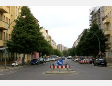 Straßenszene in einer Wohngegend mit mehrstöckigen Gebäuden, teils von Bäumen gesäumt; im Vordergrund eine Verkehrsinsel mit Verkehrsschildern und Absperrungen, geparkte Autos an den Straßenrändern. Im Hintergrund sind größere Wohngebäude zu sehen.