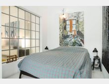 Schlafzimmer mit einem Doppelbett, das von einer blauen Tagesdecke mit rosafarbenem Muster bedeckt ist, flankiert von zwei schwarzen Nachttischen mit Lampen. Im Hintergrund eine Glaswand mit quadratischem Rahmen und ein großformatiges Gemälde an der weißen Wand.