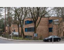 Backsteingebäude mit flachen Dächern und blauen Fensterrahmen, umgeben von Bäumen. Im Vordergrund sind Straßenschilder mit den Beschriftungen „Fohlenweg“ und „Bussardsteig“, sowie ein schwarzes Auto am Straßenrand zu sehen.