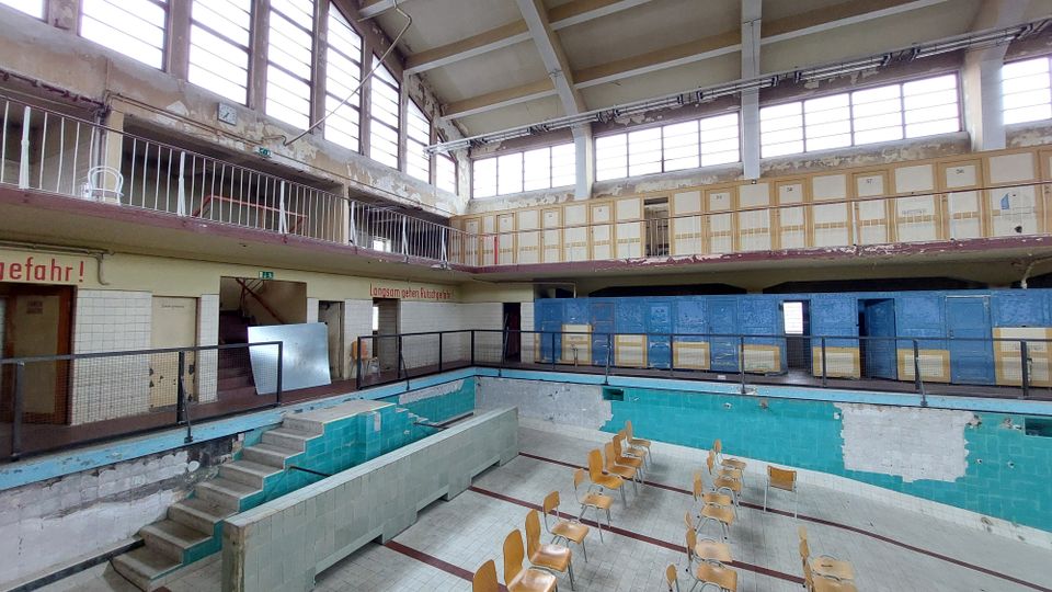 Ehemaliges Hallenbad mit leerem, verfallenem Schwimmbecken. Die Wände sind teils mit blauen und türkisfarbenen Fliesen verkleidet, umgeben von Umkleidekabinen und einer Galerie mit Geländer; im Becken stehen Stühle.