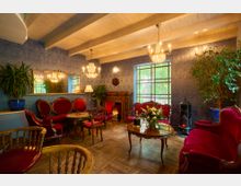 Gemütlicher Salon mit roten Polstermöbeln, Holztischen und dekorativem Kamin. Die Wände sind mit blauer Muster-Tapete gestaltet, durch große Fenster fällt Tageslicht, und Kronleuchter sowie Pflanzen sorgen für eine elegante Atmosphäre.