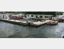 Steg mit festgemachten Booten und einem größeren Hausboot mit der Aufschrift „Schiffskontor“ auf einem ruhigen Gewässer, umgeben von Bäumen und weiteren Booten im Hintergrund.