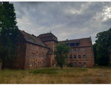 Backsteinbau mit mehreren Stockwerken, leicht baufällig, umgeben von hoher, verwilderter Wiese und großen Bäumen; dunkle Wolken bedecken den Himmel.
