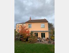 Ein zweigeschossiges Einfamilienhaus mit orangefarbener Fassade und rotem Ziegeldach, umgeben von einem Garten mit herbstlichem Laub. Vor dem Haus befindet sich eine überdachte Terrasse mit Gartenmöbeln.