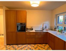 Moderne Küche mit L-förmigen Holzschränken, einem eingebauten Backofen, einem Kochfeld und einer Spüle. Die Arbeitsfläche ist weiß, der Boden hat ein geometrisches Muster in warmen Farben, und ein großes Fenster sorgt für Tageslicht.