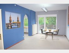 Wohnzimmer mit blau gestrichener Wand, an der ein großes gerahmtes Bild der Oberbaumbrücke hängt. Der Raum enthält einen kleinen Esstisch mit zwei schwarzen Stühlen, eine große Fensterfront mit Blick ins Grüne und einen Heizkörper.