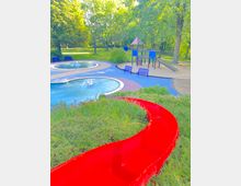 Spielplatz in einer Parklandschaft mit viel Grün und hohen Bäumen. Im Vordergrund eine rote Rutsche, im Hintergrund ein Klettergerüst mit Rutsche, Bänke und zwei runde Wasserspiele mit Springbrunnen.