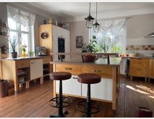 Helle, gemütliche Wohnküche mit Holzboden, Küchenmöbeln in Holz- und Weißtönen und einer zentralen Kücheninsel mit Holzplatte. Große Fenster mit weißen Vorhängen lassen Tageslicht herein, ergänzt durch Topfpflanzen und dekorative Details.