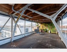 Überdachte Dachterrasse mit Stahlträgerkonstruktion, Holzbalkendecke und Gitterrostboden, umgeben von großen Glasfronten mit Blick auf umliegende Gebäude. In einer Ecke befindet sich eine begrünte Pflanzenwand mit Topfpflanzen.