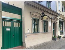 Straßenansicht eines historischen Gebäudes mit beiger Fassade, grünen Fenstern und einem grünen Tor auf der linken Seite. Rechts hängt ein Schild mit der Aufschrift „Kosmetik“ über einer Eingangstür, die ebenfalls grün gestrichen ist.