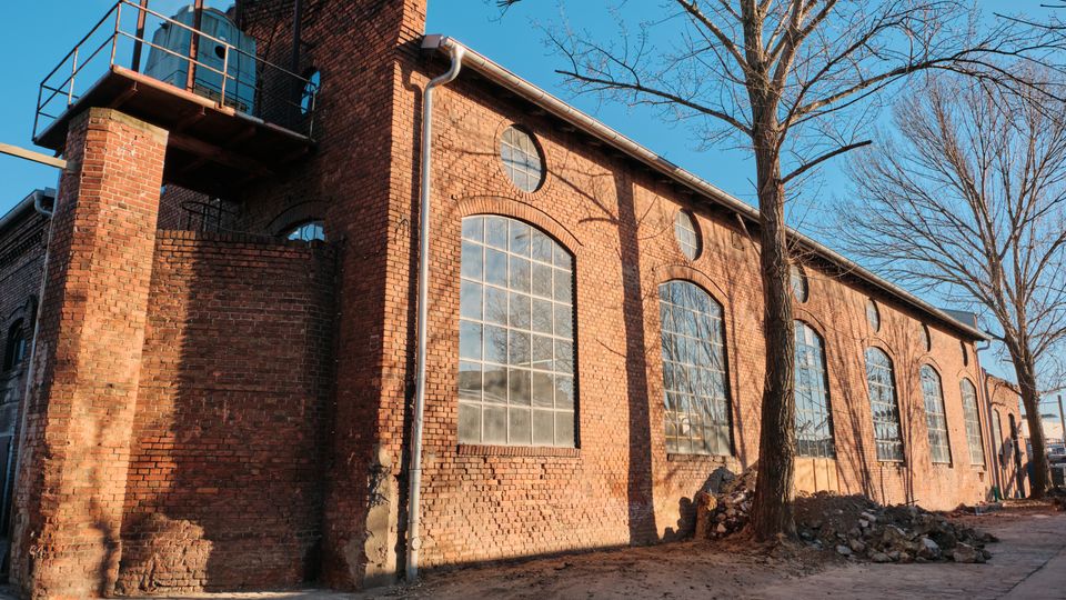 Exterior of a long red-brick industrial building with tall arched multi-pane windows and small circular windows beneath the roofline. A metal platform and water tank sit on a brick tower at the corner, with leafless trees and a paved walkway alongside the building.