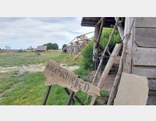 Holzgebäude und Freilichtgelände mit einer Wiese, auf der ein hölzernes Schild mit der Aufschrift „Bogenschießen“ steht; im Hintergrund sind einfache Holzhütten und eine offene Konstruktion erkennbar.