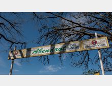 Ein Schild mit der Aufschrift „Kolonie Abendrot – Zur Kantine“, flankiert von Werbelogos der Biermarke „Berliner Kindl“, vor einem blauen Himmel mit kahlen Baumästen im Hintergrund.