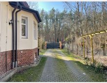 Seitenansicht eines Hauses mit weißer Fassade und Backsteinsockel, daneben ein gepflasterter Weg, der zu einem schwarzen Tor mit Backsteinsäulen führt. Rechts des Weges befinden sich ein Metallzaun und ein kleines hölzernes Rankgerüst, dahinter Wald mit kahlen und nadeligen Bäumen.