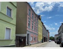 Gepflasterte Straße in einer Wohngegend mit mehrstöckigen Häusern in unterschiedlichen Farben, darunter Grün, Rosa und Gelb. Die Gebäude haben klassische Fassadenelemente wie Gesimse und verzierte Fenster, während parkende Autos am Straßenrand zu sehen sind; der Himmel ist blau mit wenigen Wolken.