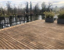 Holzterrasse mit umlaufendem schwarzen Geländer, an dem Blumentöpfe befestigt sind, und mehreren großen Pflanzkästen mit Bambus. Im Hintergrund ein Teich, umgeben von Bäumen, sowie vereinzelte Gebäude.