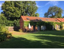 Backsteingebäude mit roten Dachziegeln und begrünten Fassaden, umgeben von einem Garten mit Rasenfläche, Bäumen und einem Holzzaun. Im Vordergrund steht eine Terrasse mit Gartenmöbeln und einem Sonnenschirm.
