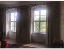 Ein schlicht eingerichteter Raum mit zwei großen, durch weiße Vorhänge gesäumten Fenstern, die in einen grünen Außenbereich blicken. Die Wände wirken rau und alt, ein einfacher Holzboden sowie ein roter Sessel und ein kleiner Beistelltisch sind zu sehen.