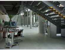 Moderner, industriell gestalteter Innenraum mit Betonwänden und -säulen, einer breiten Metalltreppe mit gelb-schwarzem Markierungsstreifen und mehreren iMac-Computern auf kleinen Schreibtischen. Der Raum hat einen offenen, minimalistischen Charakter.